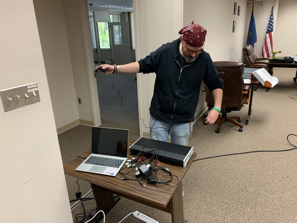 A man wearing a black hoodie and a red bandana stands next to a table set up for technical work, with a laptop and tangled cables. He appears to be organizing or connecting equipment in an office environment.