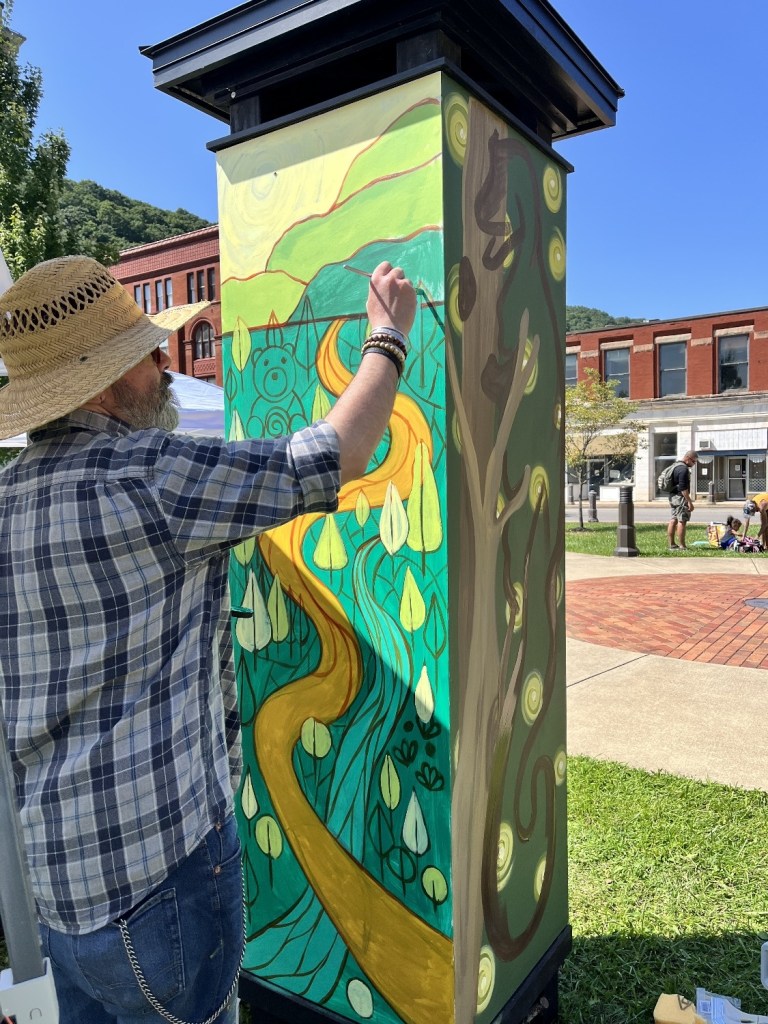 An artist painting a colorful mural on a tall structure outdoors, featuring a winding road, green hills, and abstract tree designs. The artist is wearing a straw hat and has a leisurely expression as they work.