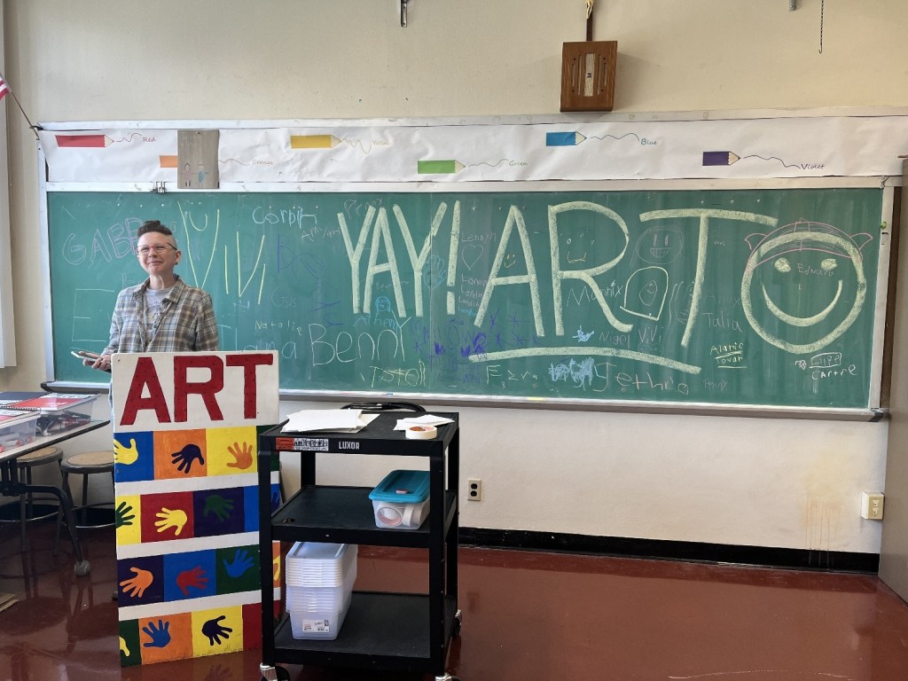 A smiling person stands next to a colorful 'ART' display in a classroom filled with chalk drawings on a green chalkboard. The chalkboard features the words 'YAY ART' and various doodles, while the background shows art supplies and classroom furniture.