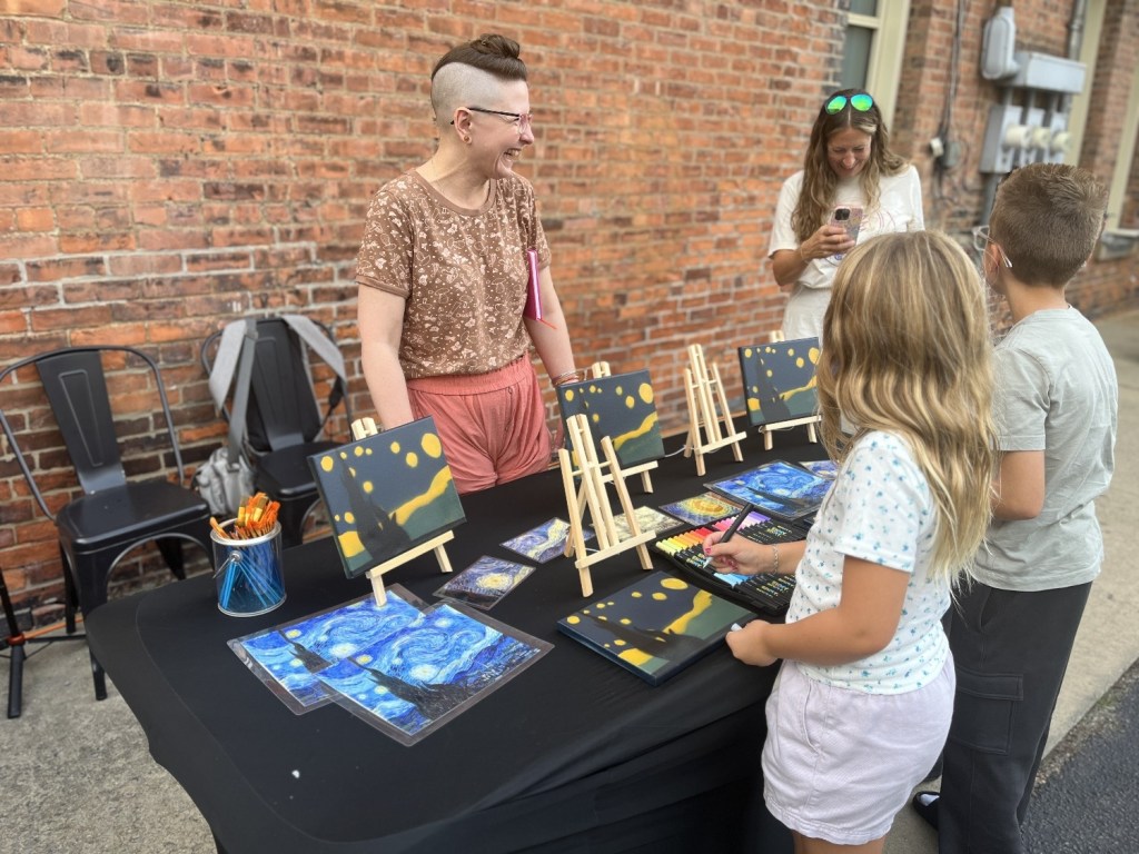 An artist at a table displaying paintings and supplies, interacting with two children who are drawing. Another adult is taking a photo in the background.
