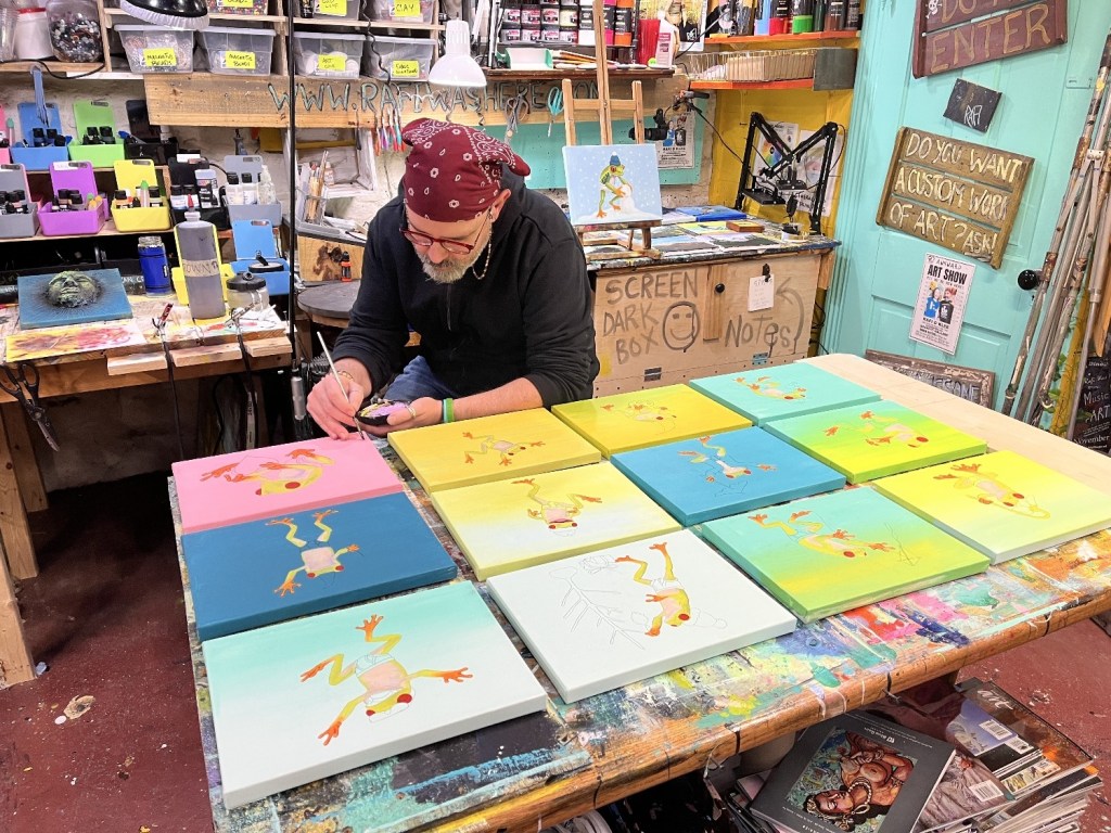 An artist wearing a headband works on colorful frog paintings arranged on a table in a vibrant studio filled with art supplies.