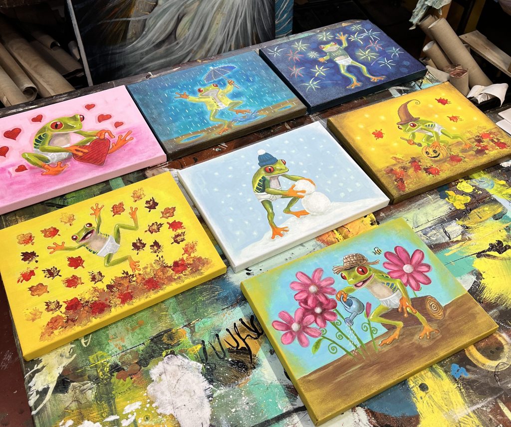 A collection of colorful paintings featuring cartoon frogs in various seasonal settings, each engaging in different activities like holding hearts, playing in leaves, rolling a snowball, and watering flowers. The paintings are arranged on a table with a vibrant and textured background.
