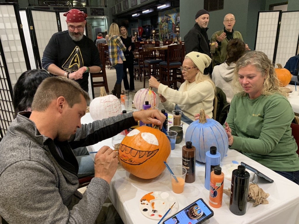 Participants engaged in a pumpkin painting event, with various painted and unpainted pumpkins on the tables while others socialize in the background.
