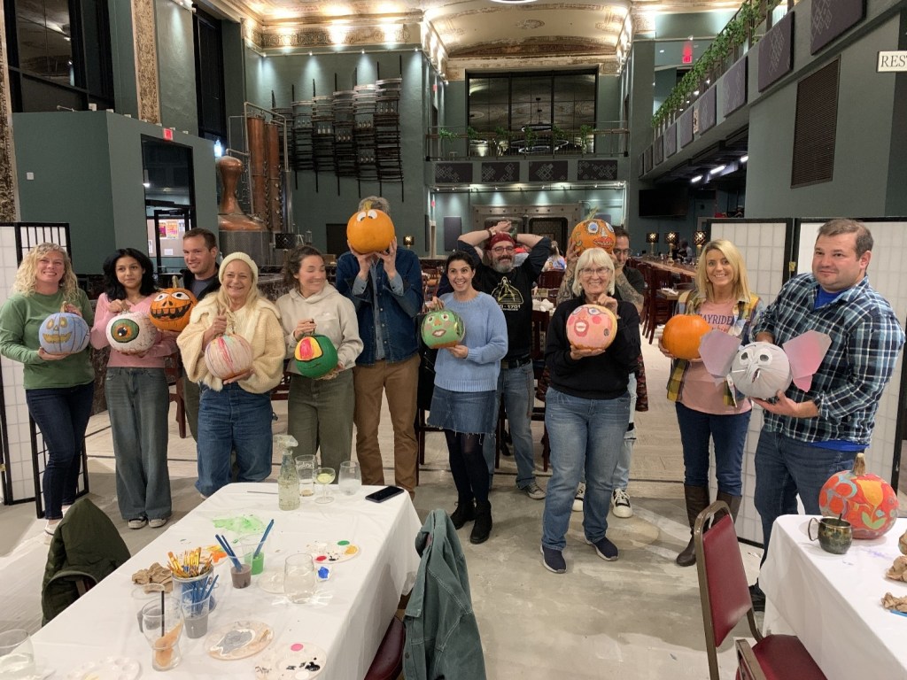 A group of people standing together in a festive setting, each holding uniquely painted pumpkins. The scene appears to be part of a pumpkin painting event, with tables covered in art supplies.