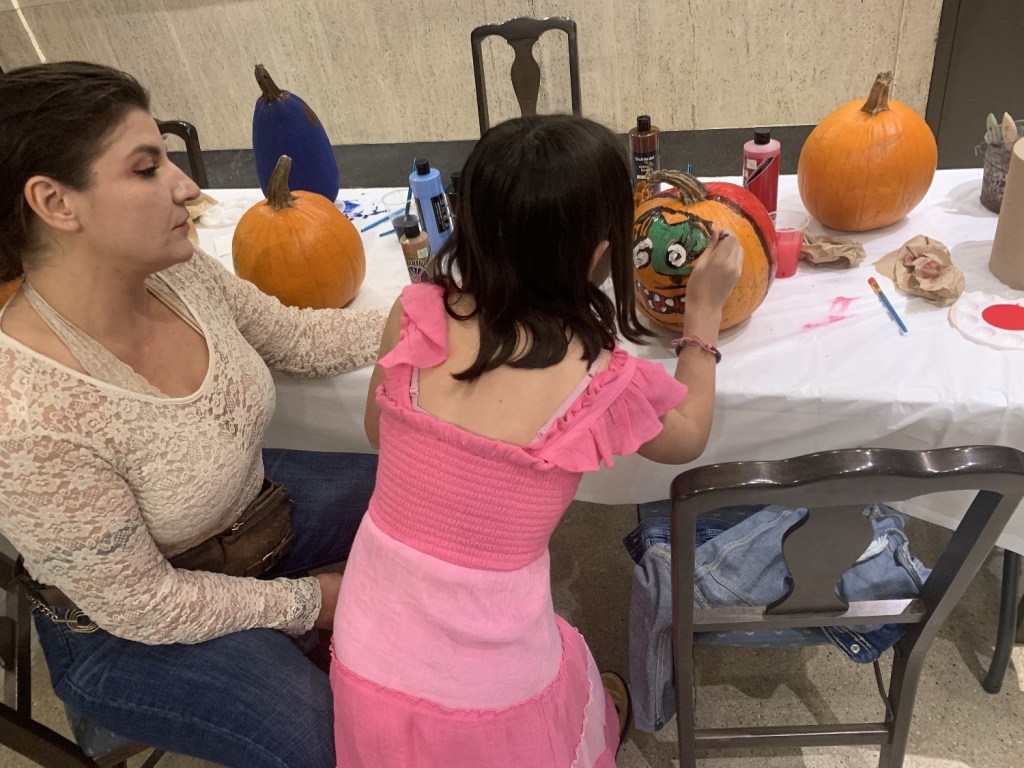 A woman and a girl painting pumpkins at a table. The woman is watching as the girl decorates a pumpkin with a colorful design. Several painted pumpkins and art supplies are visible on the table.