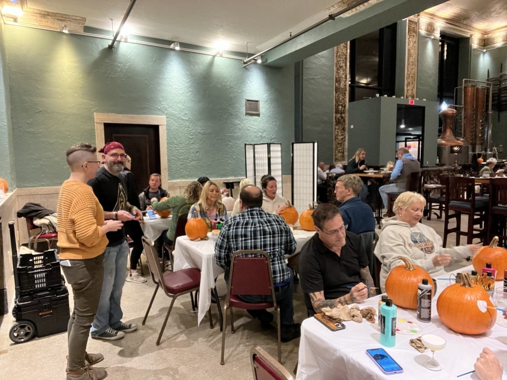 A lively indoor gathering with people seated around tables, painting and decorating pumpkins. The decor features a green wall and warm lighting, creating a cozy atmosphere.